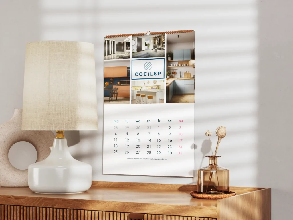 Calendario de pared con diseño minimalista y fotografías de cocinas modernas de la marca Cocilep, colocado sobre un mueble de madera junto a una lámpara decorativa.